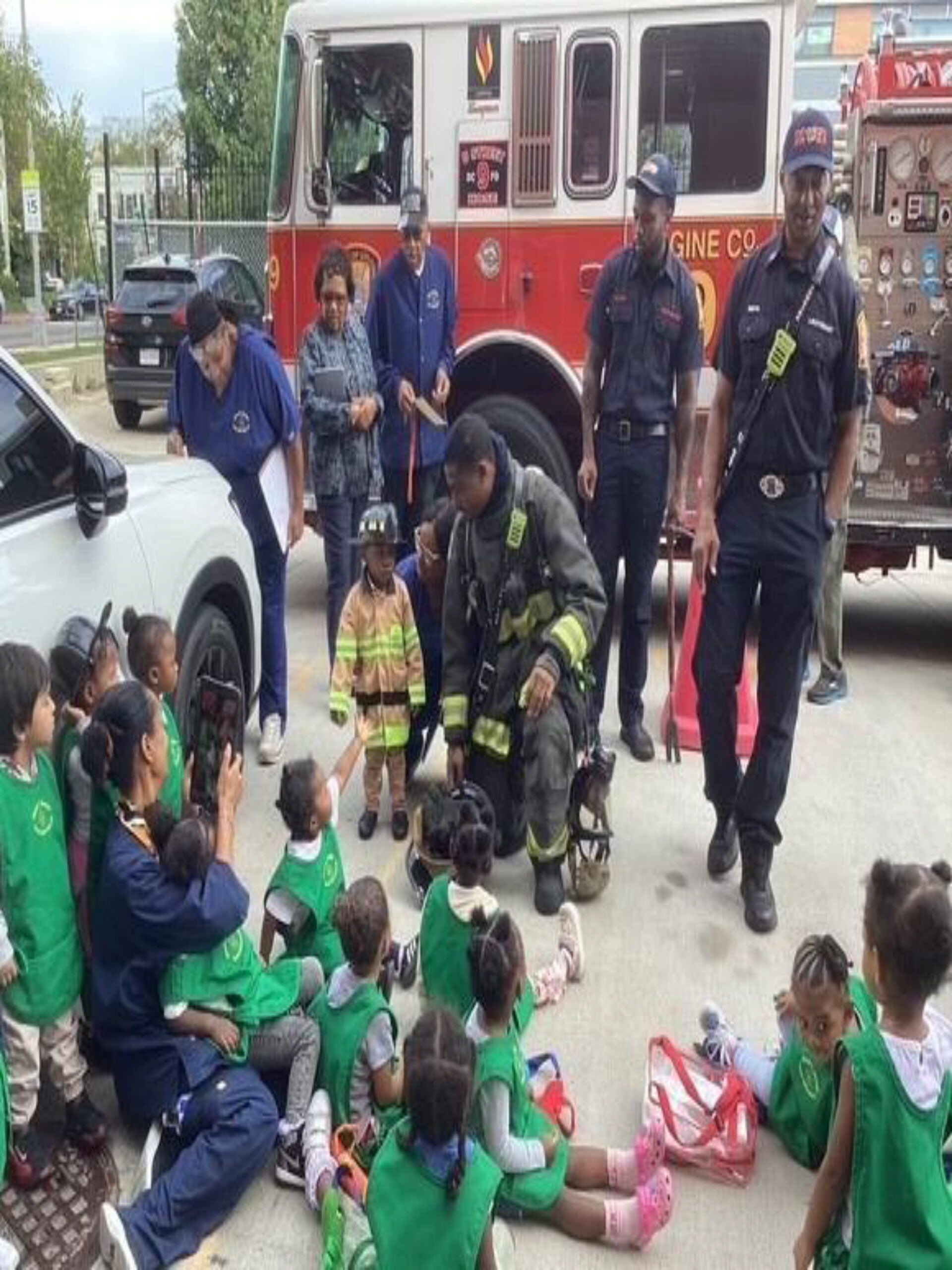 Children dressed in green vests interact with firefighters and a fire truck during a safety demonstration