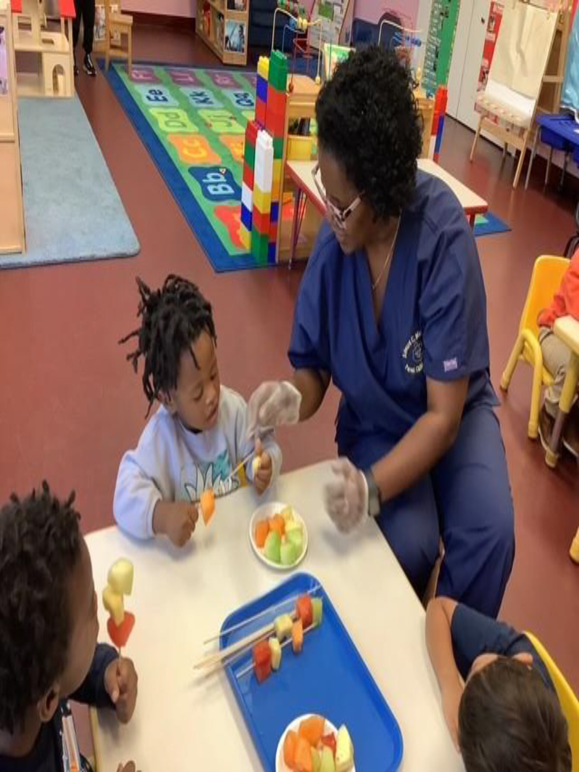 A caregiver helps children prepare fruit skewers in a colorful classroom