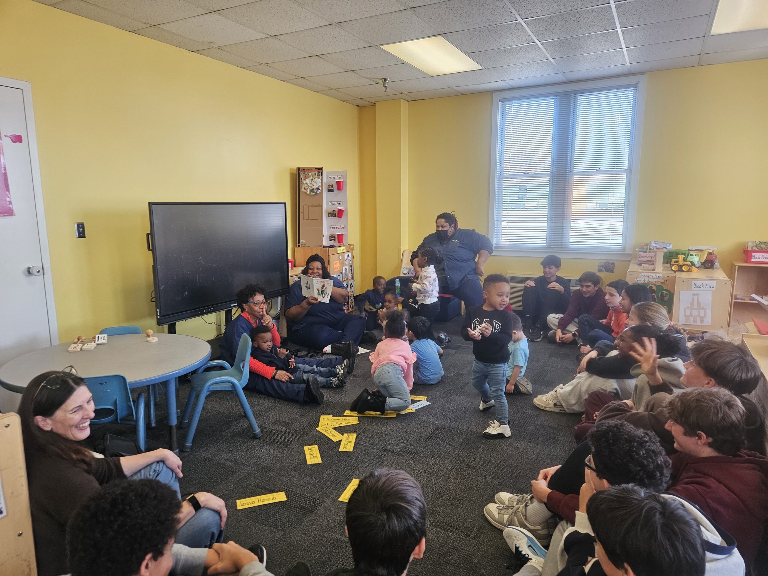 Students, teachers, and young children gathered in a classroom during a group reading and learning activity.
